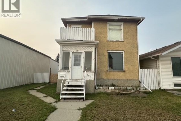 Front exterior view of a two-story yellow house with white trim and a small balcony. - 310 High Street W, Moose Jaw
