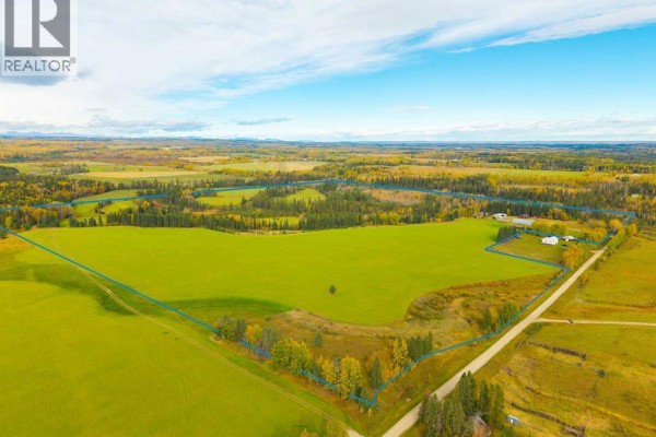 Aerial view of a large, fenced green field with scattered trees and a farmstead in the distance. - 31064 Range Road 52, Rural Mountain View County