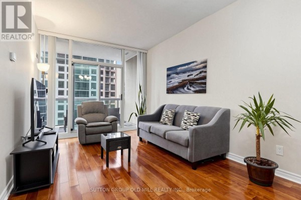 A living room with a grey sofa, armchair, hardwood floors, and a balcony view. - 3109 - 3504 Hurontario Street, Mississauga