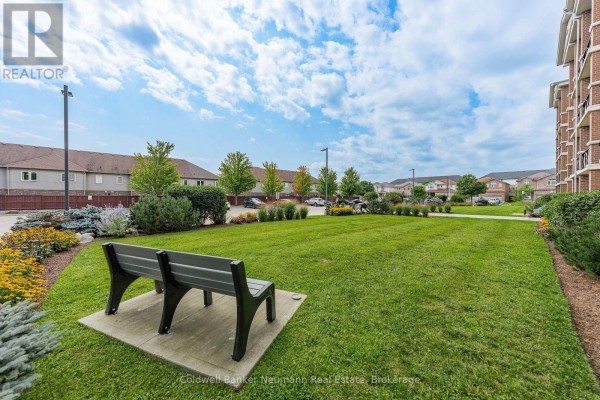Outdoor common area with a green lawn, flower beds, and a grey park bench. - 311 - 2 Colonial Drive, Guelph