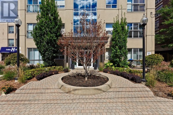 Close-up of the building entrance with a circular paved walkway and decorative tree. - 311 - 4198 Dundas Street W, Toronto