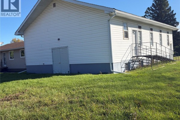 Side exterior of a white vinyl sided building with multiple windows and a metal ramp leading to a door. - 311 & 208 Main Street, Pelly