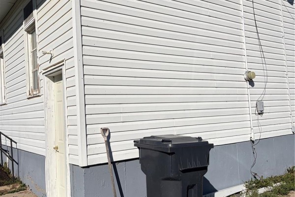 Exterior view of a white vinyl sided building corner showing a side door, trash bin, and shovel. - 311 & 208 Main Street, Pelly