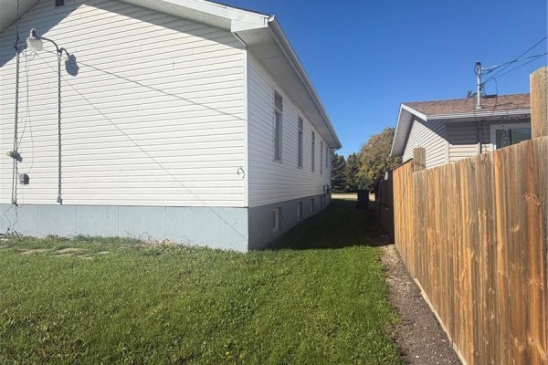 Side exterior of a white vinyl sided building with a wooden fence on the right and grassy lawn. - 311 & 208 Main Street, Pelly