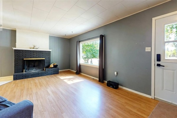 Living room with a black brick fireplace, grey walls, light wood floors, and a white door. - 311 Pottery Street, Eastend