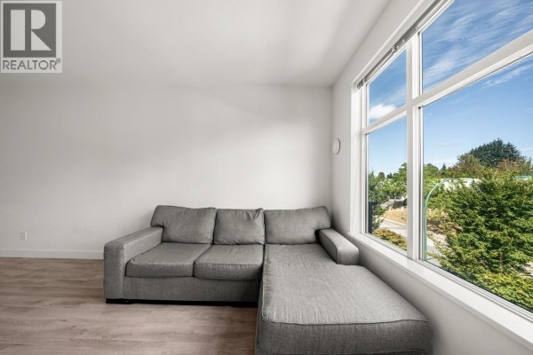 Living room corner with a grey sectional sofa, large window showing green trees and a bright blue sky. - 312 828 Gauthier Avenue, Coquitlam