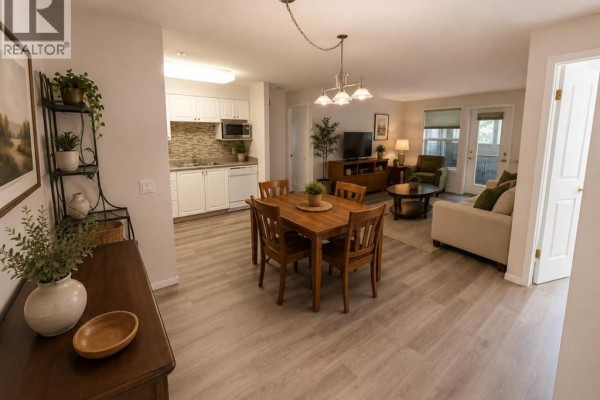 Open-concept living and dining area with a kitchen, wood dining table, sofa, and a view of the patio. - 3120, 3000 Millrise Point Sw, Calgary