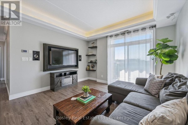 Living room featuring a large television mounted on the wall, a dark grey sectional sofa, and a large window with sheer curtains. - 314 - 2 Colonial Drive, Guelph