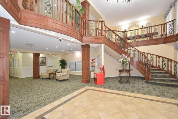 Grand lobby with a sweeping wooden staircase, patterned carpet, and tile flooring. - #314 9820 165 St Nw, Edmonton