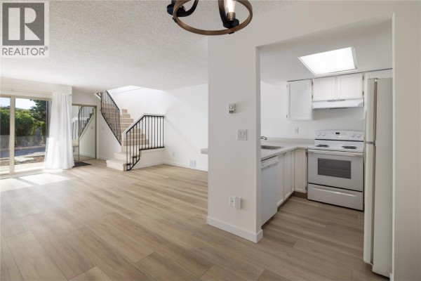 Living room and kitchen with white appliances, light wood-look flooring, and stairs. - 3140 Wilson Street Unit# 916, Penticton