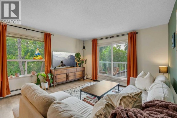 Living room with large windows, balcony access, sectional sofa, and wood-look flooring. - 317, 2211 29 Street Sw, Calgary