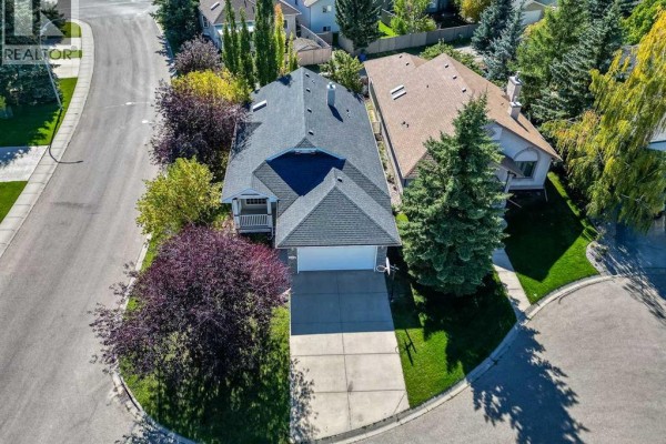Aerial view of a beige house with a dark roof, situated on a corner lot with mature trees and green lawn. - 319 Cimarron Bay, Okotoks