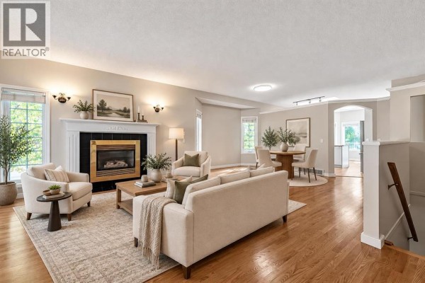 Living room with a fireplace, seating area, dining table, and hardwood floors, with an archway leading to another room. - 319 Cimarron Bay, Okotoks