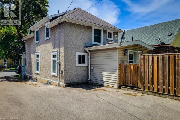 Rear exterior of a house with light-colored siding, a wooden fence, and a paved area. - 319 King Street W, Dundas