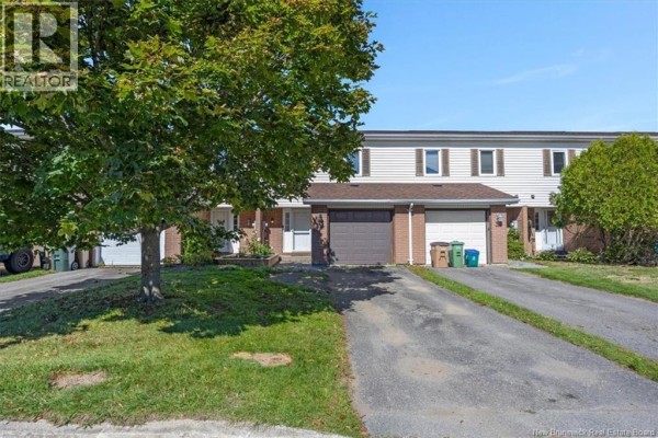 Street view of townhouse units with garages, a mature tree in the foreground, and green spaces. - 32 Driftwood Lane, Saint John