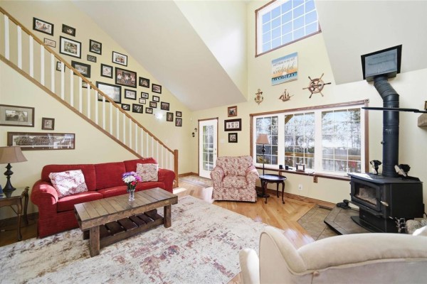 Living room with red sofa, armchair, wood stove, and a staircase with framed pictures. - 32 Eastshore Drive, Victoria Beach
