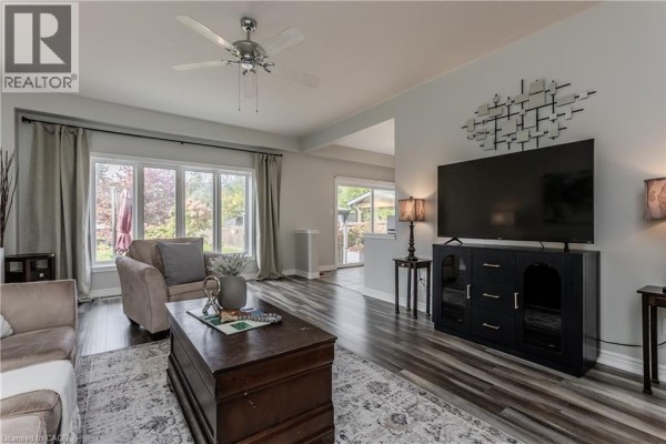 Living room with a large television on a dark media console, beige sofa, armchair, and windows overlooking the backyard. - 32 Great Oak Trail, Binbrook