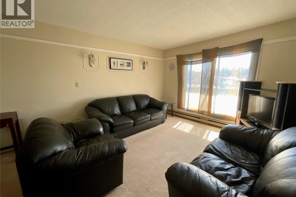 Living room with black leather sofas, beige carpet, and large windows with sheer curtains. - 320 Northgate Unit# 211, Tumbler Ridge