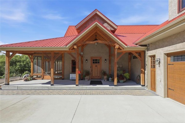 Covered front porch with wooden beams, a wooden front door, and surrounding landscaping. - 32022 Rat River Road, St Pierre Jolys