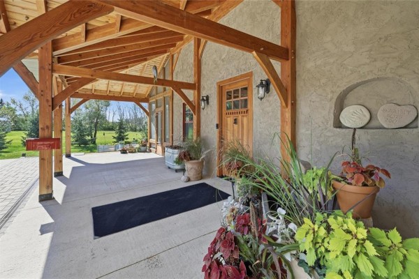 Close-up of the covered front porch entrance with large wooden beams and potted plants. - 32022 Rat River Road, St Pierre Jolys