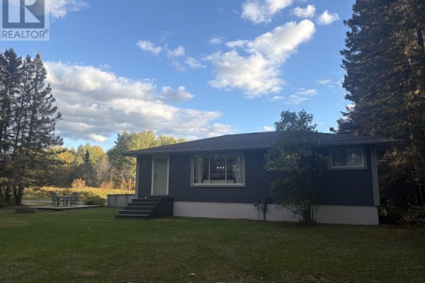 Side view of a dark grey bungalow with a lawn and deck in front, backed by trees and a field. - 3203 Hwy 61 Highway, Thunder Bay Remote Area