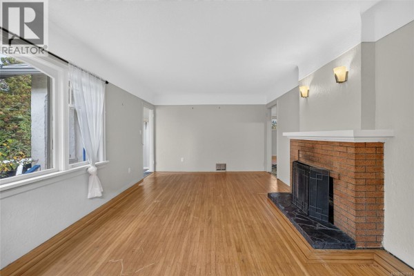 Spacious living room with hardwood floors, brick fireplace, and large windows on two walls. - 3219 Rutledge St, Saanich