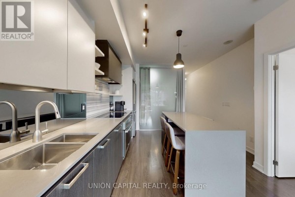 Long kitchen with double sink, grey cabinets, white countertops, and a kitchen island with bar stools. - 323 - 1830 Bloor Street W, Toronto