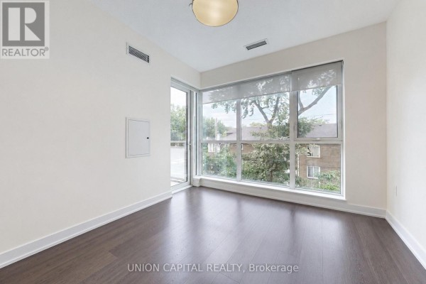 Empty room with dark hardwood floors and a large corner window with blinds and city views. - 323 - 1830 Bloor Street W, Toronto