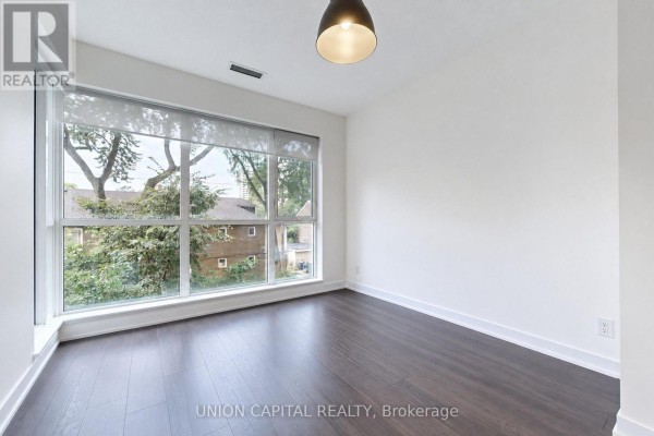 Empty room with dark hardwood floors and a large window with blinds and greenery view. - 323 - 1830 Bloor Street W, Toronto