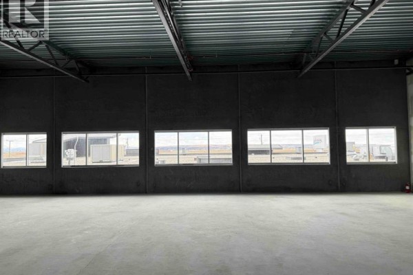 Empty commercial space with concrete floor, dark grey walls, and multiple windows facing industrial buildings. - 3235, 6520 36 Street Ne, Calgary