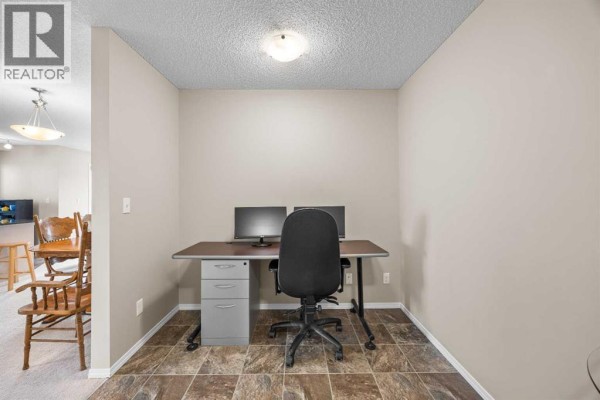 Home office area with beige walls, tiled floor, desk, two monitors, and an office chair. - 324, 195 Kincora Glen Road Nw, Calgary