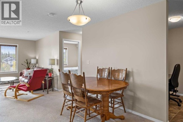 Living and dining area with beige walls, carpeted floors, red chairs, and a wooden dining set. - 324, 195 Kincora Glen Road Nw, Calgary