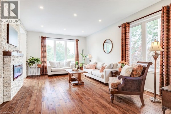 Living room with white sofas, a wooden coffee table, a fireplace, and large windows. - 3244 Steeplechase Drive, Burlington