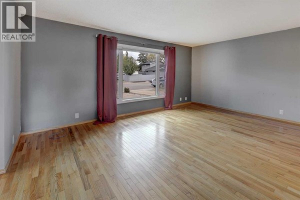 Empty living room with grey walls, hardwood floors, and large windows with maroon curtains. - 325 Flett Drive Ne, Airdrie