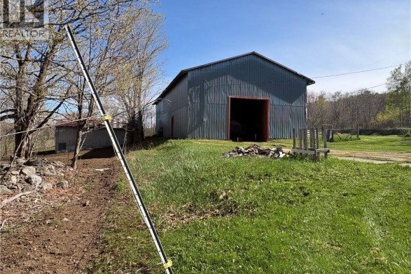 Exterior view of a large grey metal barn with a wide entrance, featuring a fence and farm equipment. - 326 Dorn Ridge Road, Burtts Corner