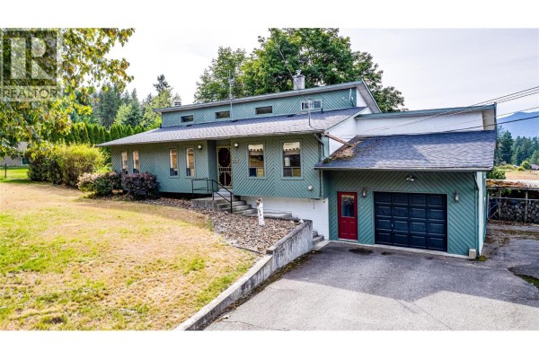 Side exterior view of a green house with a sloped driveway and a red door on the garage. - 3275 Upper Mcleod Road, Spallumcheen