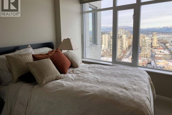 Bedroom with a large window overlooking the city, a bed with decorative pillows, and beige walls. - 32xx 888 Carnarvon St, New Westminster