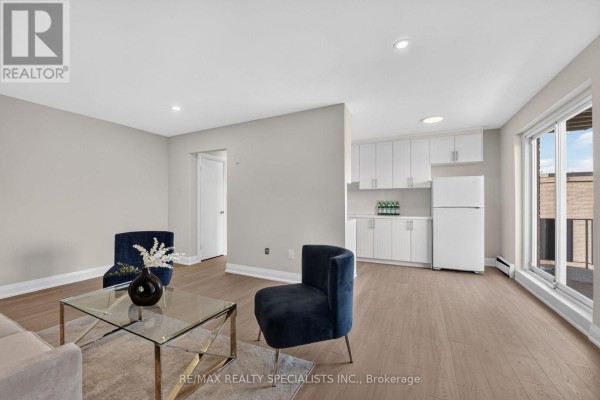 Living room and kitchen area with light grey walls, light wood floors, white cabinets, and a large window. - 33 - 426 Queenston Street, St  Catharines