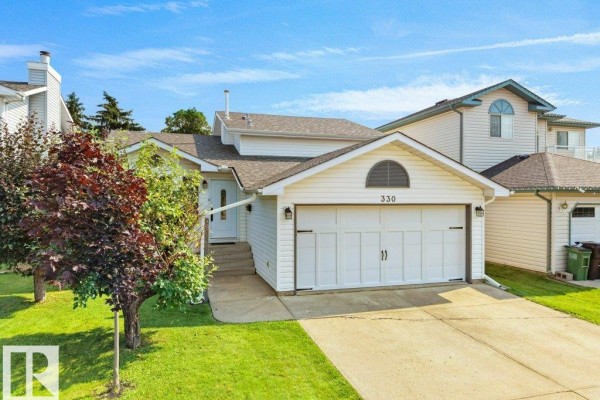 Exterior view of a white stucco house with a double garage, a manicured lawn, and mature trees. - 330 Pearson Cr Nw, Edmonton