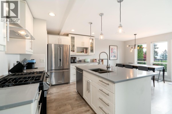 View of a modern white kitchen with stainless steel appliances, a gray island with sink, and a dining area beyond. - 330 Richmond Street, New Westminster