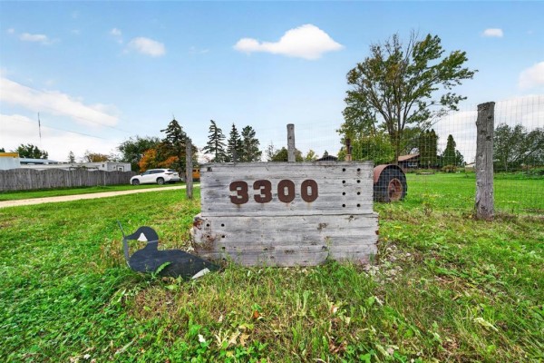Wooden sign with large carved numbers '3300' sits in a grassy area with a fence and an old tractor. - 3300 St Mary's Road, Winnipeg