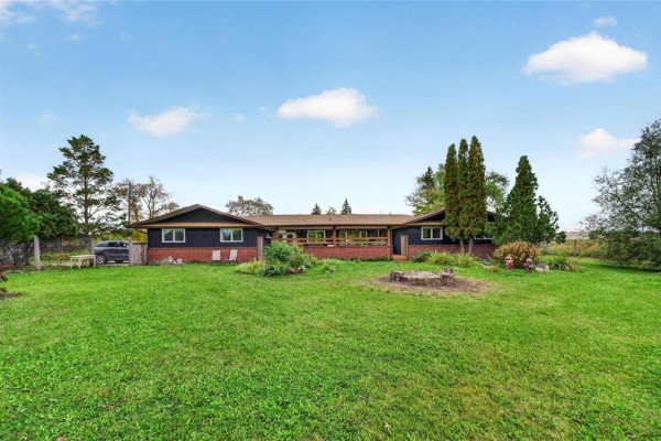 Front view of a sprawling, dark brown house with a brick base, featuring a wide wooden deck and fire pit. - 3300 St Mary's Road, Winnipeg