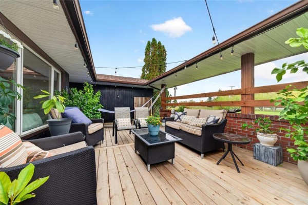 Wooden deck with outdoor furniture, string lights, and potted plants, adjacent to a dark wood-sided house. - 3300 St Mary's Road, Winnipeg