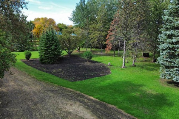 Green lawn with manicured beds and mature trees on a sunny day, viewed from a gravel driveway. - 33153 25 Road W, Carman