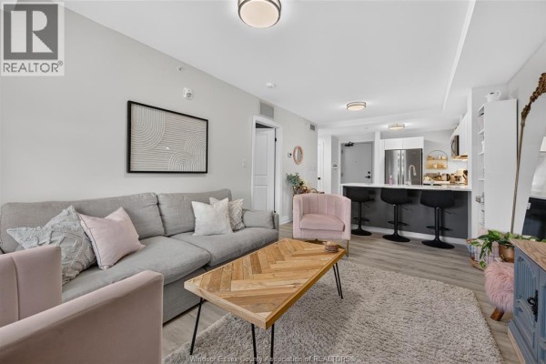 Open concept living room and kitchen area with grey sectional sofa, pink chairs, and dark grey island. - 3320 Stella Crescent Unit# 109, Windsor