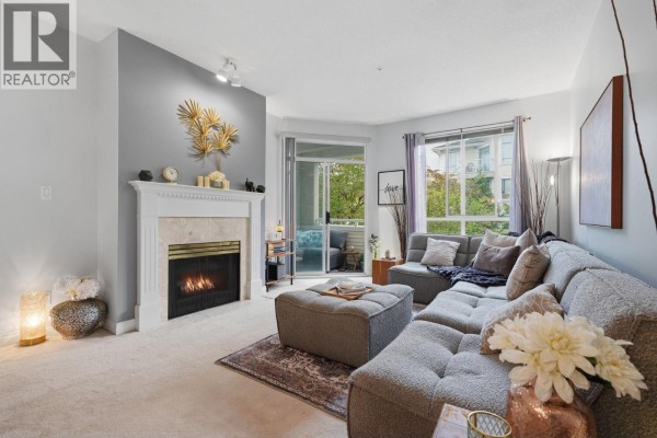 Living room with a fireplace, large sectional sofa, ottoman, and window view of trees. - 333 383 E 37th Avenue, Vancouver