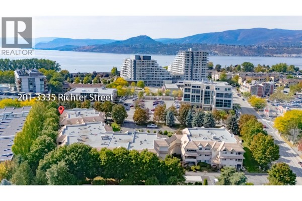 Aerial view of an apartment complex with surrounding trees and a distant view of the lake and mountains. - 3335 Richter Street Unit# 201, Kelowna