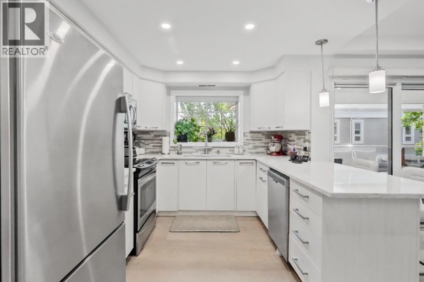 Modern white kitchen with stainless steel appliances, a double sink, and a window overlooking greenery. - 3335 Richter Street Unit# 201, Kelowna