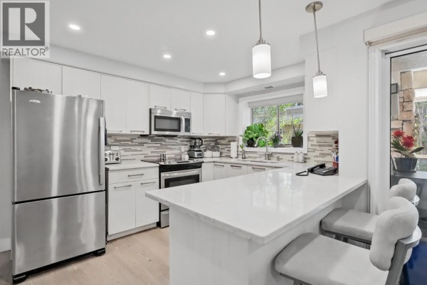 Kitchen island with quartz countertop and seating, adjacent to white cabinetry and stainless steel appliances. - 3335 Richter Street Unit# 201, Kelowna