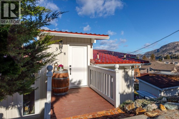 Close-up of the entrance with a wooden deck, red roof, and decorative barrel, surrounded by greenery. - 334 Carmel Crescent, Okanagan Falls